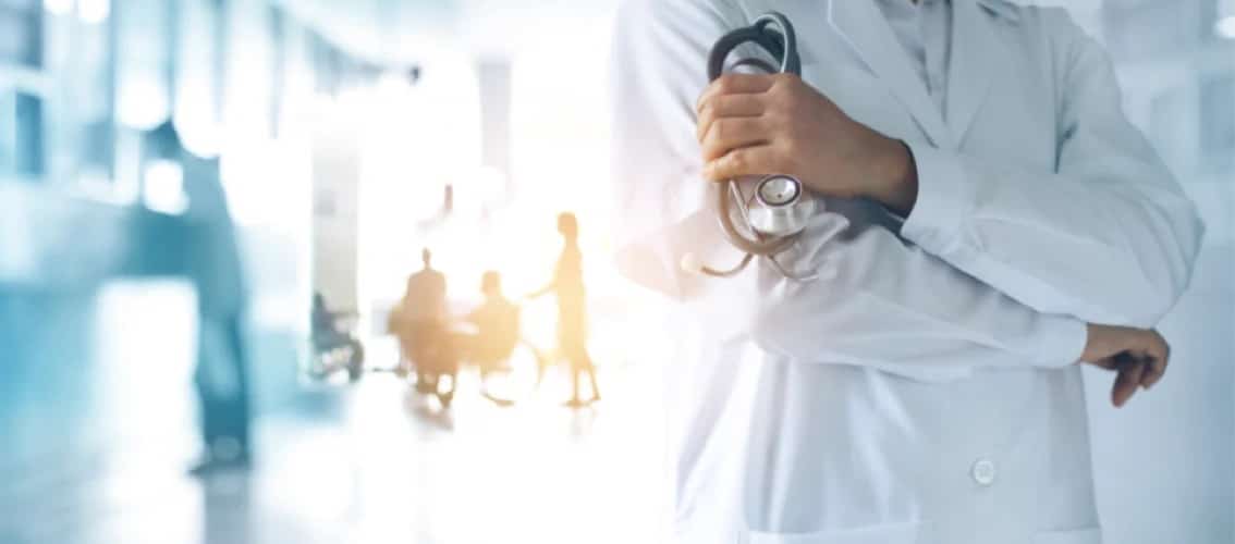 A physician in a white coat folds their arms with hospital in background
