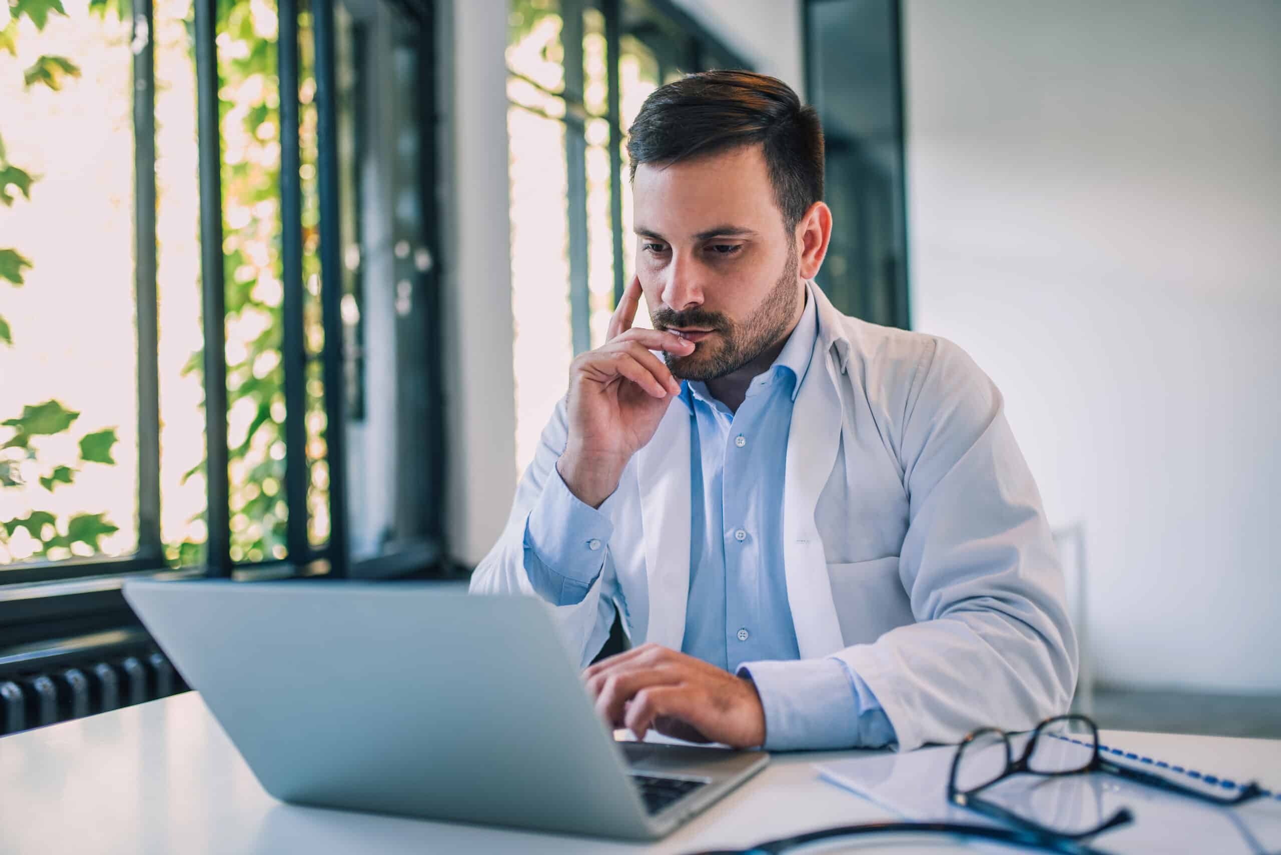 A serious male physician looks at his laptop.
