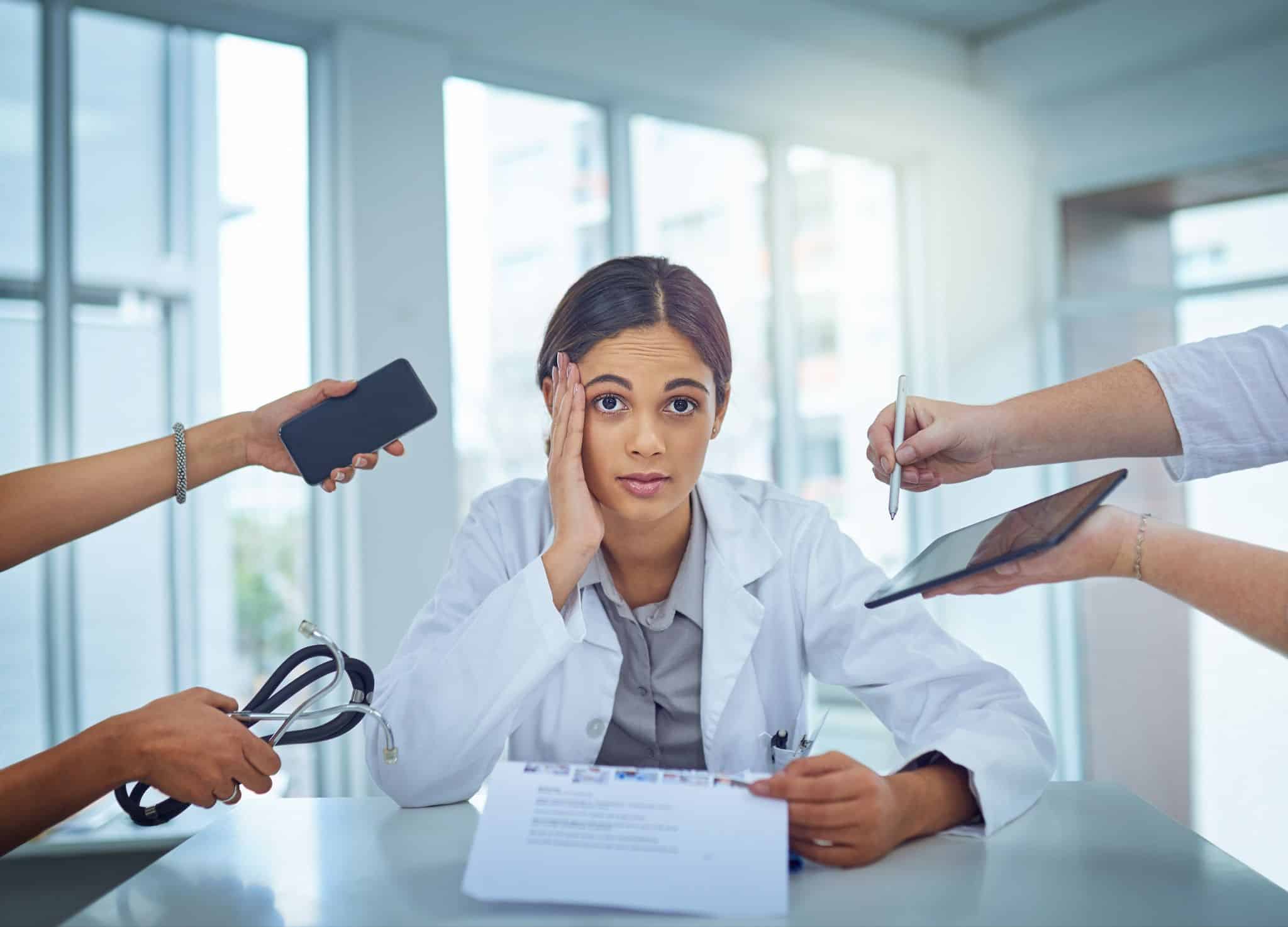 A female physician looks overwhelmed as hands try to give her a phone, tablet, pen, and stethoscope.