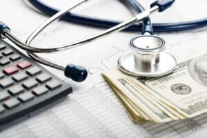 A stethoscope, calculator, and stack of $100 bills sits on top of paperwork